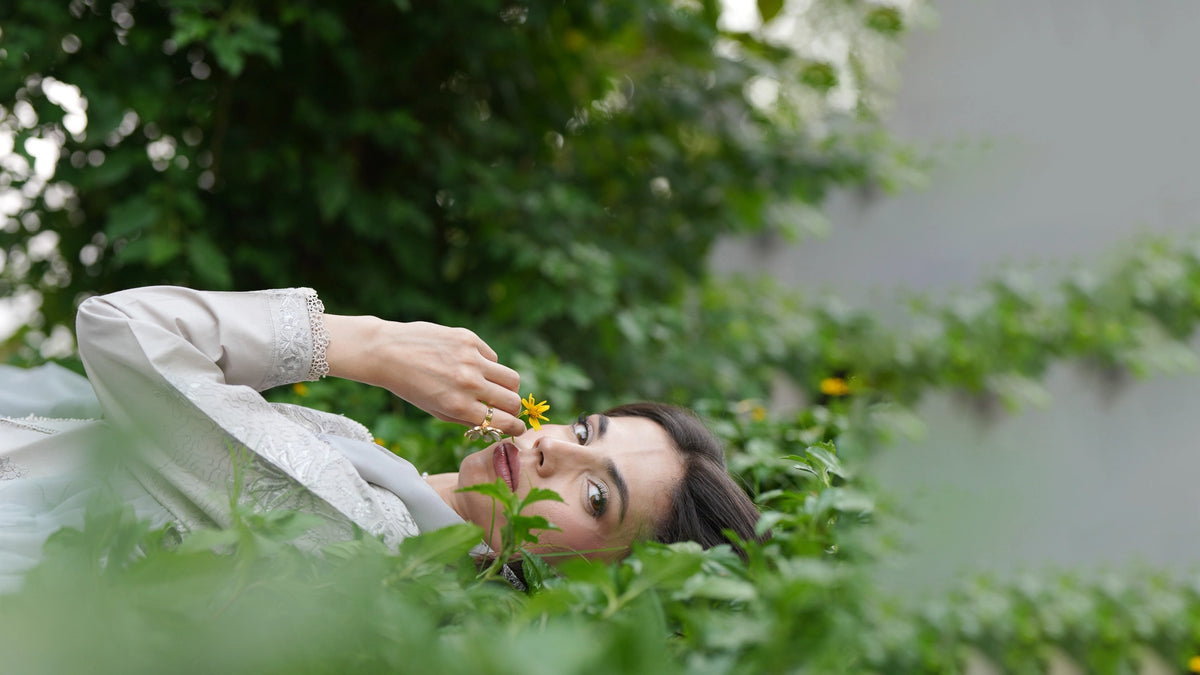 Editorial shot of a model lying among greenery holding a yellow flower, wearing a grey embroidered suit from Bad-e-Saba by INEJ.