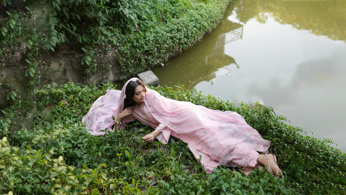 High-angle banner view of a model reclining beside water wearing a soft pink 3-piece embroidered lawn suit by INEJ.