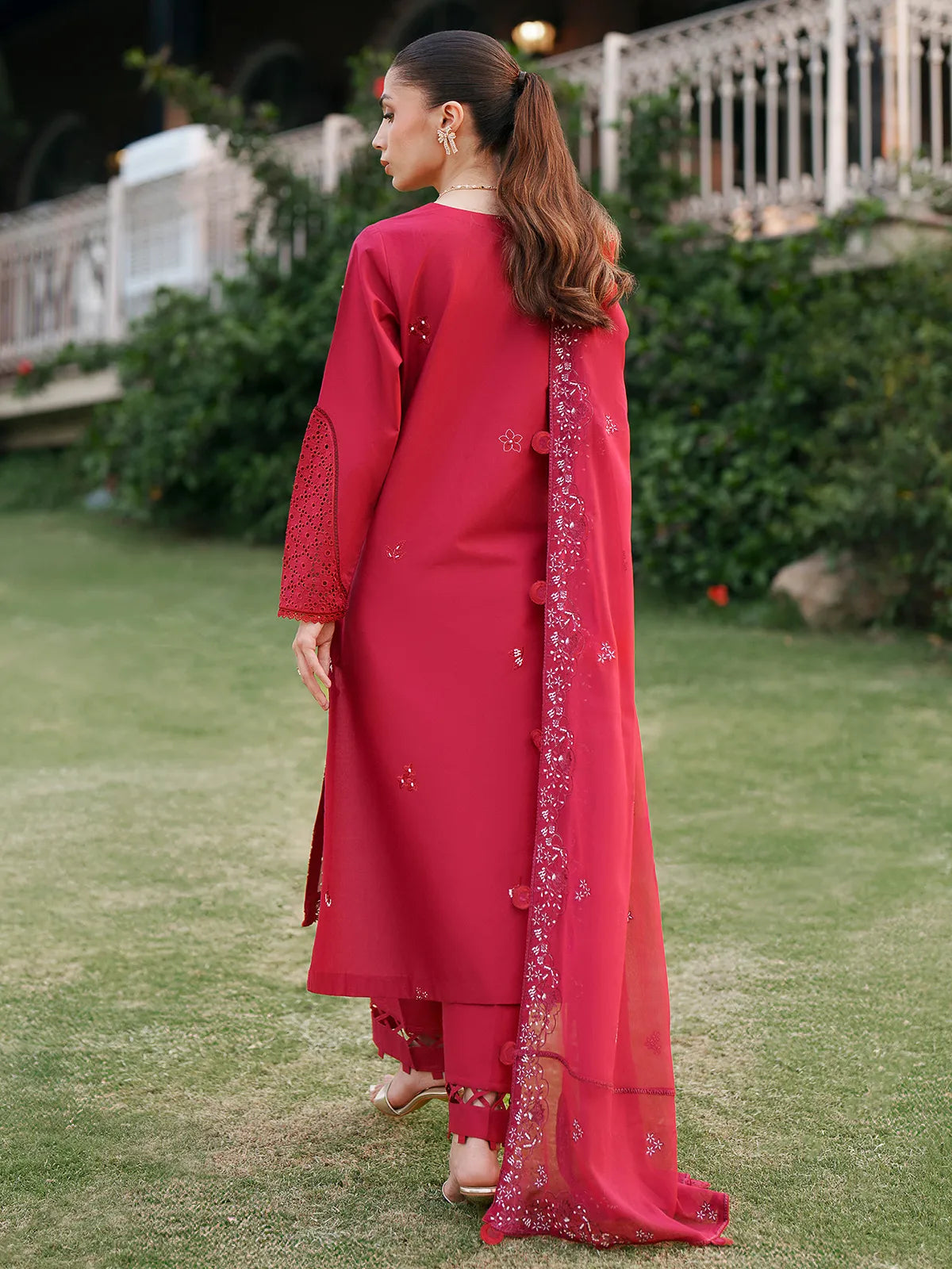 Back full-length view of vibrant red lawn stitched 3-piece suit featuring eyelet cutwork sleeves, embroidered matching dupatta draped over the shoulder, and cutwork trouser cuffs from Baad-e-Saba by INEJ.